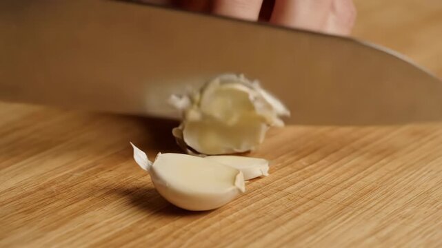 A knife smashes a garlic clove flat, exposing its texture and oils in a dramatic food prep moment