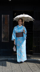 Fototapeta premium A woman in a light blue floral kimono stands at the entrance of a traditional Japanese building, holding a lace parasol and a handbag, blending elegance, heritage, and timeless cultural beauty.