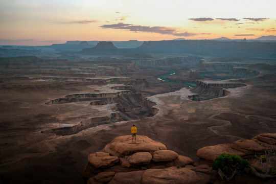 Traveler standing on the edge of Canyonlands National Park, Utah, USA, watching a breathtaking desert sunset. Vast red rock canyons, mesas, and winding Colorado River below.