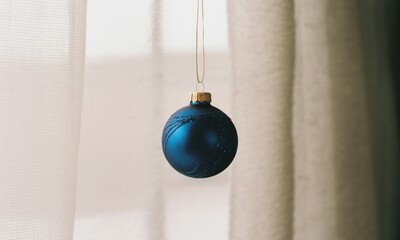 A single deep blue Christmas ornament hangs from a gold string in front of sheer white and beige curtains