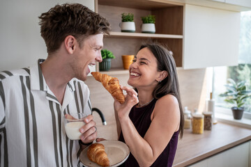 Happy couple sharing croissant and milk at breakfast in kitchen