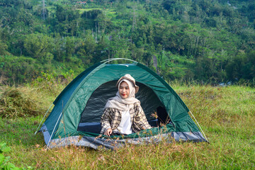 Sporty Muslim Asian woman standing near tent and backpack, enjoying sunny afternoon in the mountains. Camping lifestyle concept