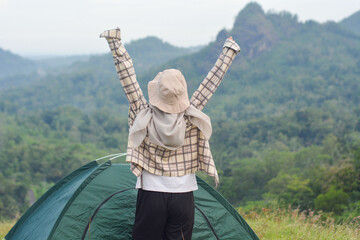 Sporty Muslim Asian woman standing near tent and backpack, enjoying sunny afternoon in the mountains. Camping lifestyle concept
