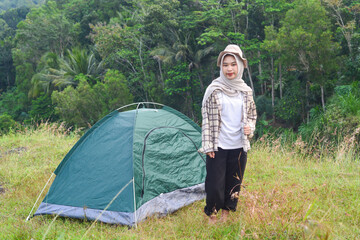 Sporty Muslim Asian woman standing near tent and backpack, enjoying sunny afternoon in the mountains. Camping lifestyle concept
