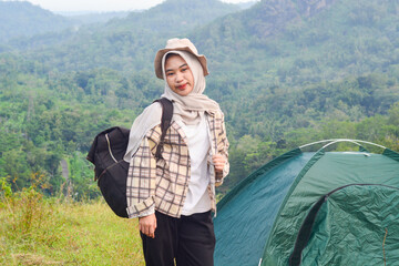 Naklejka premium Sporty Muslim Asian woman standing near tent and backpack, enjoying sunny afternoon in the mountains. Camping lifestyle concept