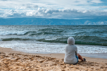 woman wearing hooded sweatshirt sits on sandy beach, observing rolling waves and mountains in...
