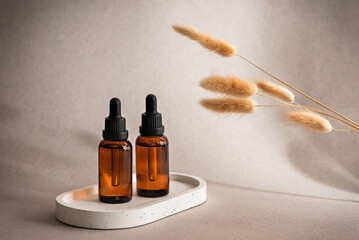 Fototapeta premium Two amber glass dropper bottles with cosmetic serum or essential oil on beige flowing background decorated with fluffy lagurus grass, selective focus, copy space, rejuvenating skin care product