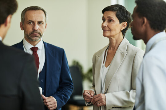 Middle aged Caucasian man and woman standing in business meeting, dressed in formal suits, listening to two colleagues, diverse group engaging in professional discussion indoors
