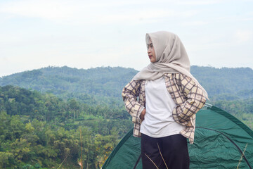 Sporty Muslim Asian woman standing near tent and backpack, enjoying sunny afternoon in the mountains. Camping lifestyle concept