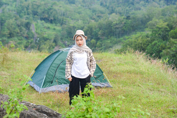 Sporty Muslim Asian woman standing near tent and backpack, enjoying sunny afternoon in the mountains. Camping lifestyle concept