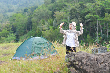 Sporty Muslim Asian woman standing near tent and backpack, enjoying sunny afternoon in the mountains. Camping lifestyle concept
