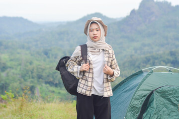 Sporty Muslim Asian woman standing near tent and backpack, enjoying sunny afternoon in the mountains. Camping lifestyle concept