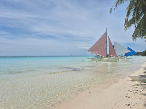 Traditional paraw sailboats with colorful sails anchored along the white sand beach of Boracay, turquoise tropical sea and clear blue sky creating a stunning paradise island seascape in the Philippine