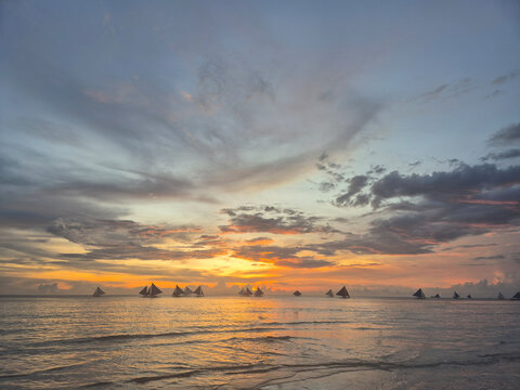Spectacular sunset seascape at Boracay White Beach Philippines with traditional paraw sailboats silhouetted on the horizon, vibrant orange purple and pink sky reflecting on calm tropical ocean water