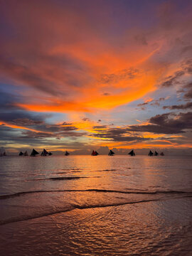 Spectacular sunset seascape at Boracay White Beach Philippines with traditional paraw sailboats silhouetted on the horizon, vibrant orange purple and pink sky reflecting on calm tropical ocean water