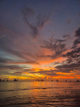 Spectacular sunset seascape at Boracay White Beach Philippines with traditional paraw sailboats silhouetted on the horizon, vibrant orange purple and pink sky reflecting on calm tropical ocean water