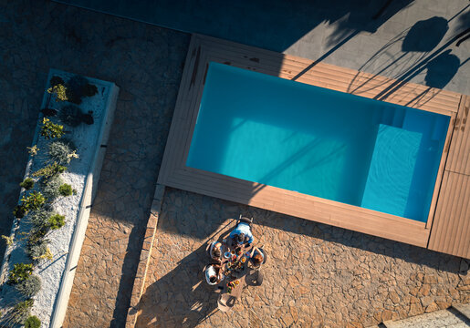 Aerial view of group of adult friends having lunch by swimming pool in sunny backyard.