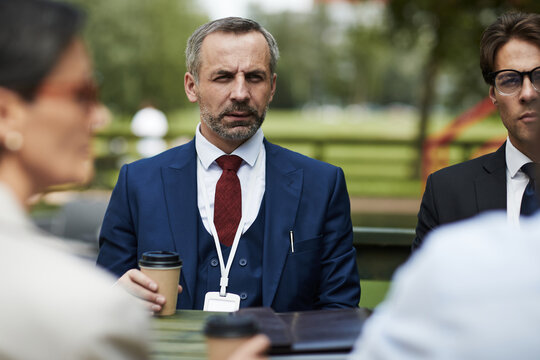 Middle aged Caucasian man with short gray hair and beard sitting outdoors at table with coffee cup, wearing suit and lanyard, surrounded by business colleagues during meeting