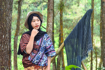 Young Asian Muslim woman hiking and camping in autumn forest, smiling at camera wearing Javanese...
