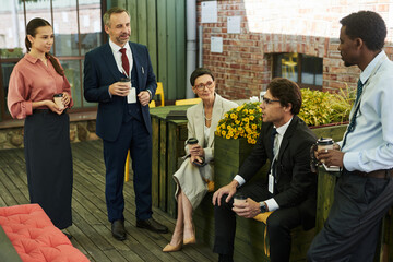 Group of middle aged multiethnic business professionals standing and sitting outdoors holding coffee cups, engaging in conversation during informal meeting on wooden terrace