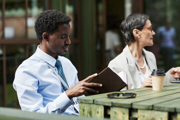 Black young businessman checking diary for appointments outdoors while sitting at wooden table next...