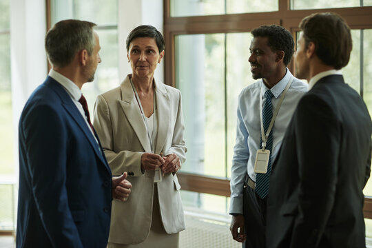 Middle aged Caucasian woman and three middle aged men, including one Black man and two Caucasian men, standing together in office environment, engaging in professional conversation