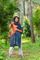 Young Asian Muslim woman hiking and camping in autumn forest, smiling at camera wearing Javanese...
