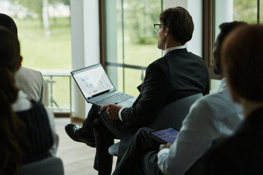 Caucasian middle aged man sitting in conference room holding laptop with business charts on screen surrounded by group of professionals listening during corporate meeting