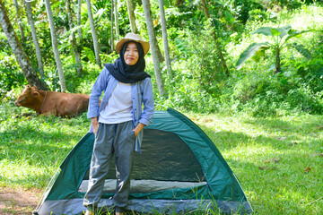 Sporty Muslim Asian woman standing near tent and backpack, enjoying sunny afternoon in the mountains. Camping lifestyle concept