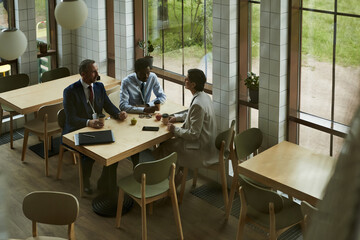 Three middle aged business people, including Caucasian, Black men and woman, sitting at table in modern cafe, discussing business while holding coffee cups and using digital tablet near large windows