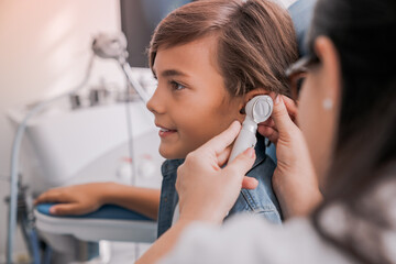 Side view shot of female doctor examining boy's ear with otoscope. Barotrauma laryngitis otitis ear treatment. Otorhinolaryngology concept
