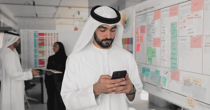 Serious Arabian professional in white Muslim dress typing on smartphone