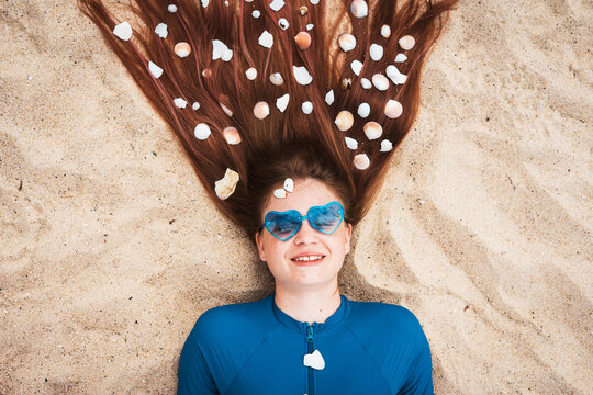 Creative summer portrait of smiling girl with seashells in her long hair, lying on sand with heart-shaped sunglasses. Concept of playful holiday vibes, beach lifestyle, and artistic freedom. - Powered by Adobe
