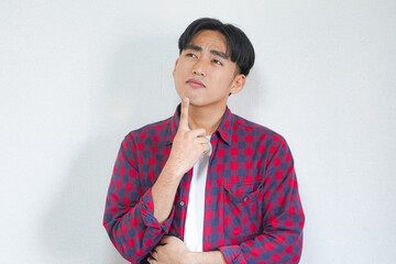 Young man in a red checkered shirt thoughtfully touches his chin, looking upwards against a white...