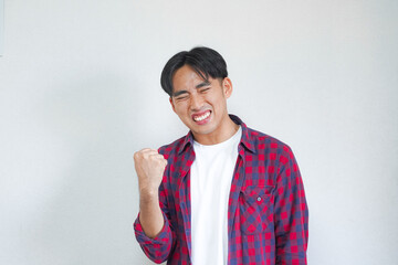 A young Asian man in a plaid shirt celebrates with a clenched fist and joyful expression against a...