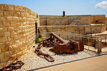 A large rusty sea anchor with a massive metal chain lies on the gravel in a historic fortress with steps and stone walls. © Elena