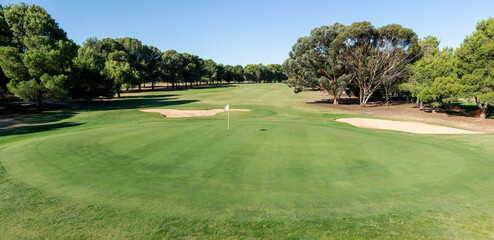 Golf course green and bunkers on a sunny day, surrounded by trees and blue sky