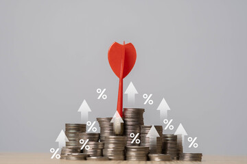 A red dart is stuck in stacked coins, surrounded by percentage symbols and upward arrows,...