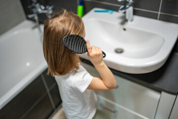 Little girl brushing her hair with a detangling hairbrush in the bathroom. Concept of daily self-care and developing independence in childhood.