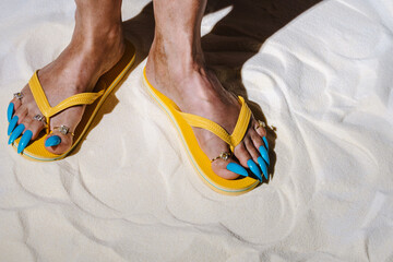 Yellow sandals and blue false nails on sand with playful summer vibe
