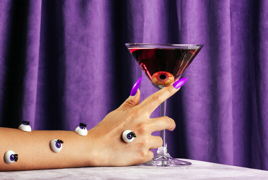 Hand with eyeball decorations holds cocktail against purple curtain