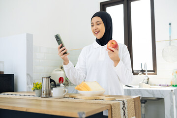 A cheerful woman in a light-filled kitchen holding an apple and checking her phone. A breakfast...