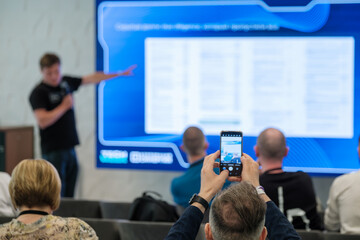 Participants in an indoor seminar recording a professional presentation on their smartphones, emphasizing visual learning and modern technologies.