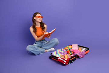 Lovely young woman sitting with an open suitcase of colorful clothes, planning a vacation in a...