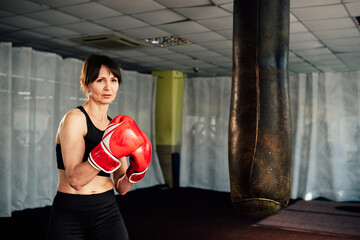 Determined mature woman wearing red boxing gloves is training with a punching bag in a gym, embodying strength, fitness, and the empowering spirit of boxing