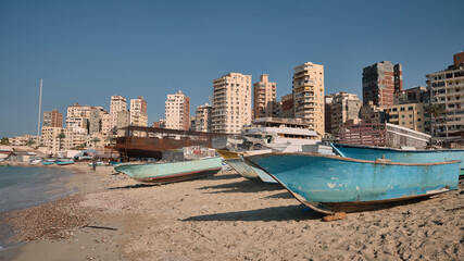 Traditional wooden fishing boats rest on sandy beach under clear blue sky. Tall apartment buildings and densely packed urban skyline rising in background