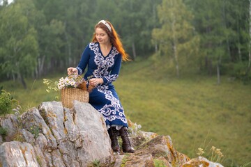 Redhead woman wearing embroidered dress sits on rocks, arranging wildflowers in basket amidst lush...