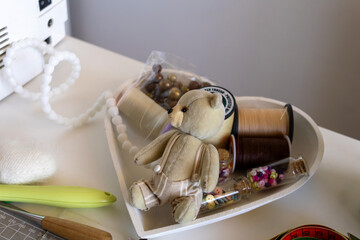 A charming handmade teddy bear sits amongst an array of crafting supplies like spools of thread,...