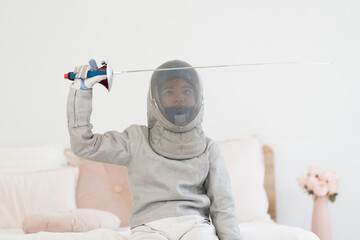 Asian girl in fencing costume with fencing sports mask and holding sword for practicing and training in gym. Teen girl fencer athlete sitting on bed during preparing for training in gym