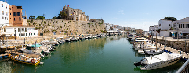Panoramic view of Mahón Harbor in Menorca, with boats docked on both sides and the fortress in the...